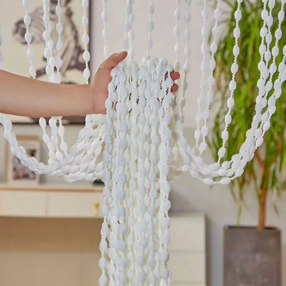A hand is pulling a white beaded string curtain with tassels, used as a bohemian door and window drape.