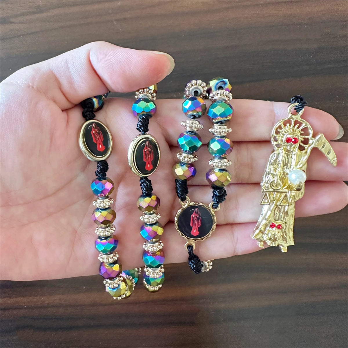 A hand holds a rosary necklace featuring black beads, green and blue gemstones, and a pendant depicting Santa Muerte.