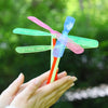 A hand holds a classic bamboo dragonfly flying propeller toy with pink and blue blades against a blurred green background.