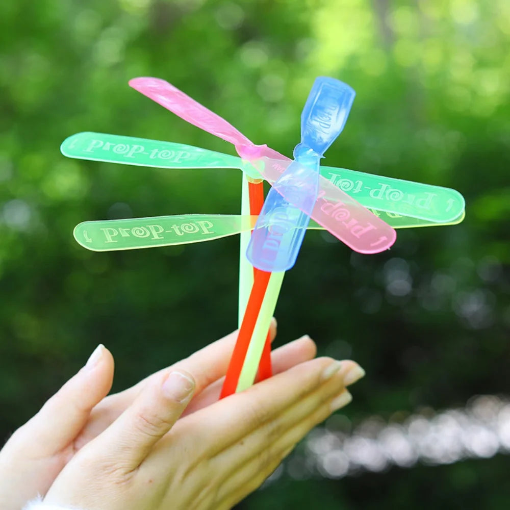 A hand holds a classic bamboo dragonfly flying propeller toy with pink and blue blades against a blurred green background.