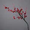 A branch of artificial plum blossoms with small red flowers and buds against a gray background.