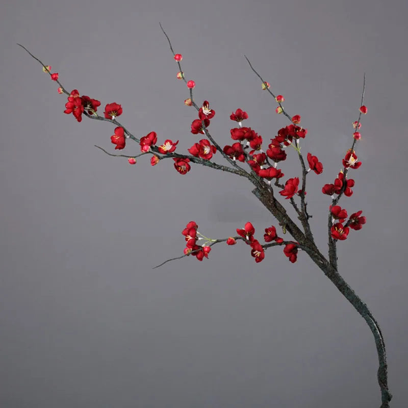 A branch of artificial plum blossoms with small red flowers and buds against a gray background.