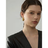 A close-up portrait of a woman wearing gold geometric stud earrings and a black garment.