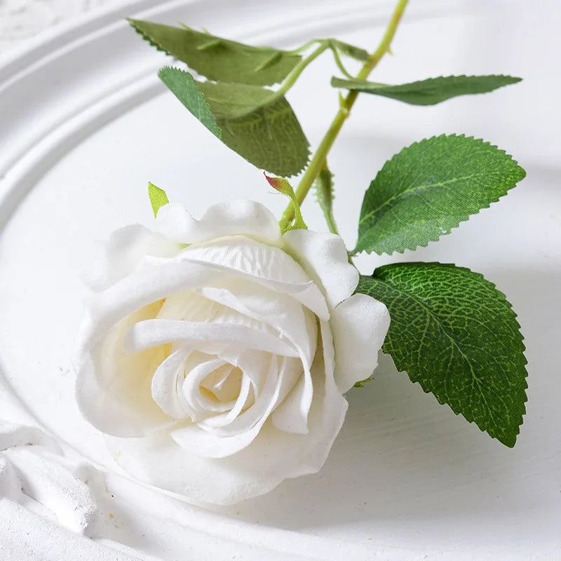 A white artificial rose with green leaves and a bendable stem placed on a white plate.