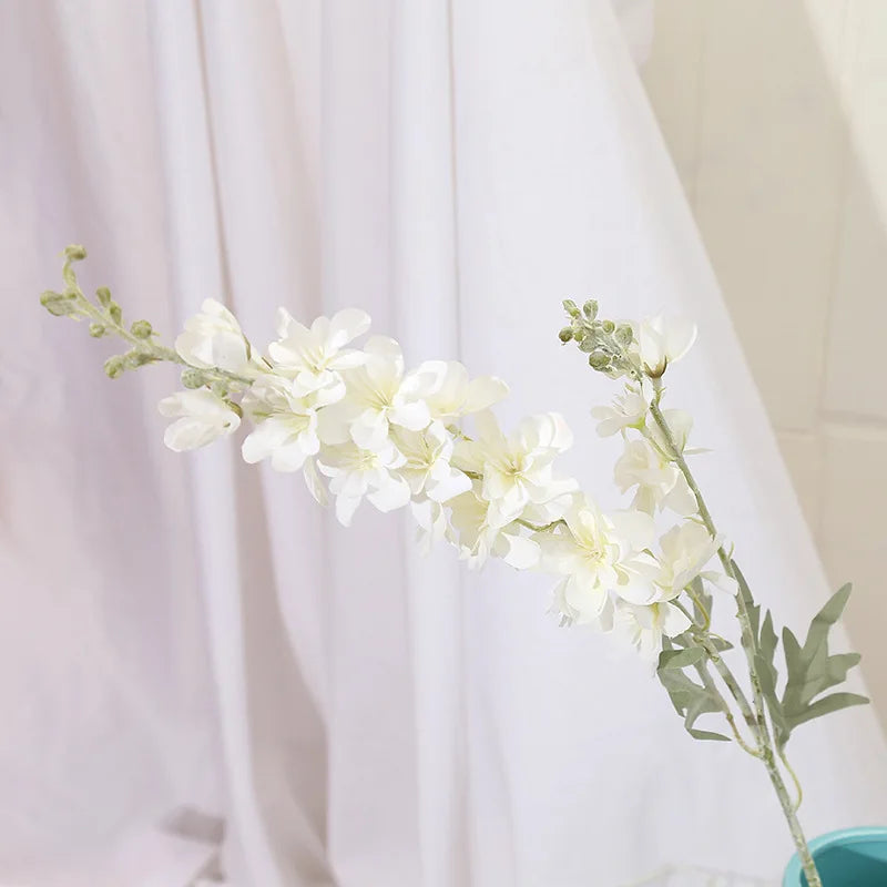 Un délicat jet de fleurs artificielles de delphinium blanches sur un fond drapé blanc.
