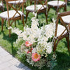 Un bouquet de fleurs artificielles de delphinium blanches et roses sur une pelouse verte à côté de chaises en bois.