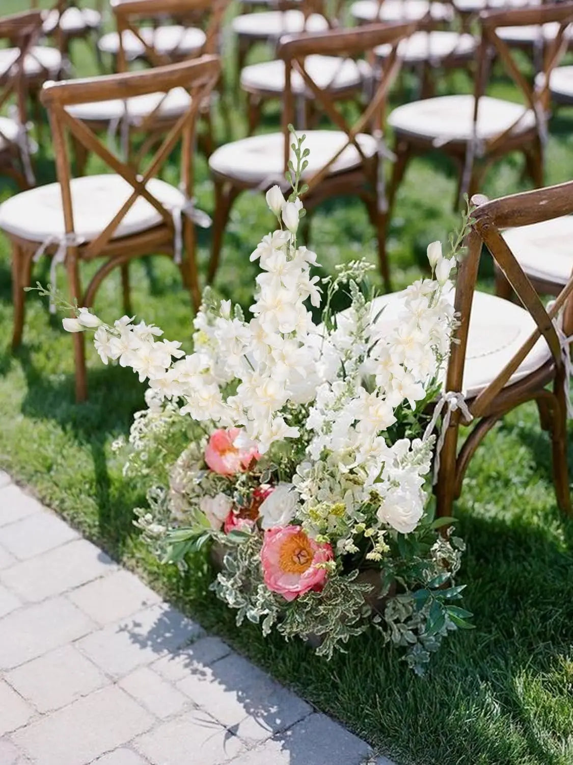 Un bouquet de fleurs artificielles de delphinium blanches et roses sur une pelouse verte à côté de chaises en bois.