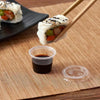A hand holds a roll of sushi wrapped in nori and topped with sesame seeds, positioned above a clear plastic sauce cup.