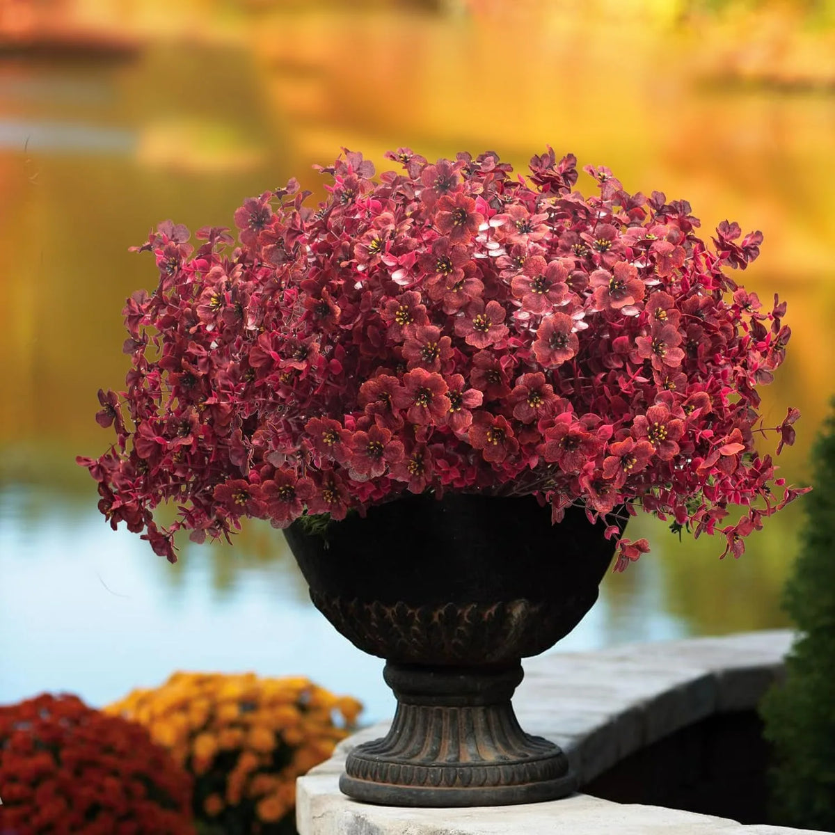 A dense mound of realistic artificial silk flowers in deep reddish-pink sits in a dark urn by water.