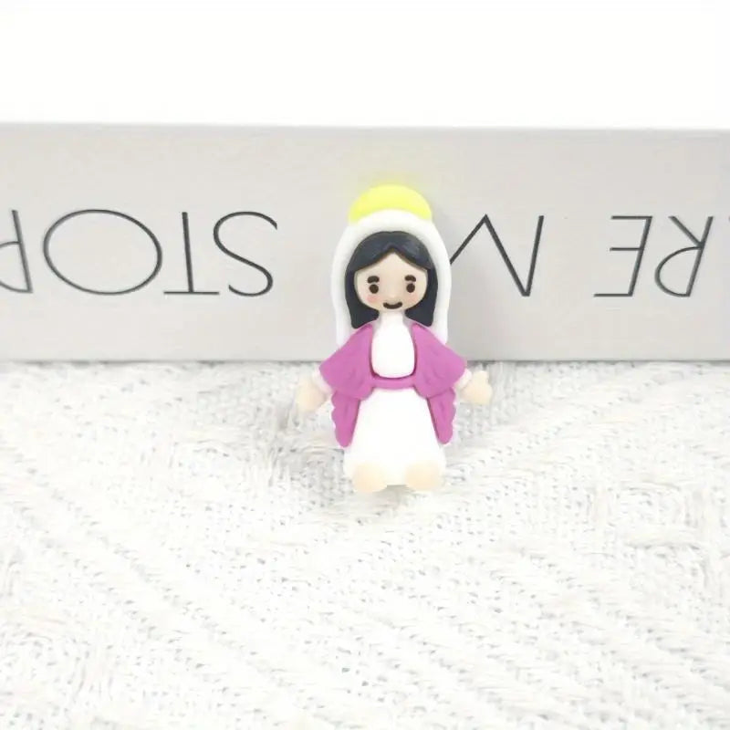 A small resin statue of the Virgin Mary with a white robe, pink garment, and yellow halo, displayed on a white textured surfa