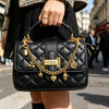 A woman holds a black quilted leather handbag with gold chain charms and a gold clasp.