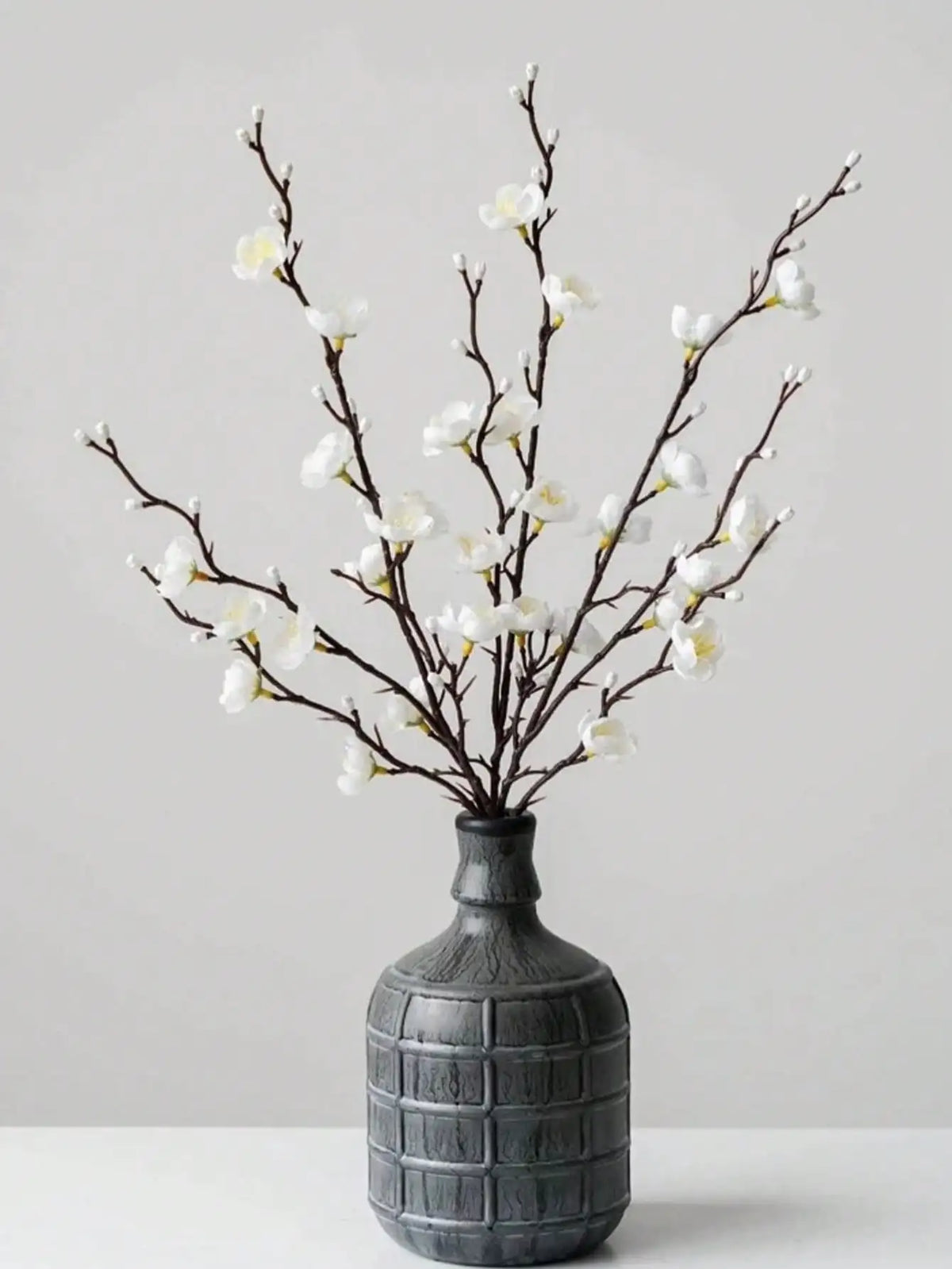 Ramitas de flor de ciruela blanca artificial dispuestas en un jarrón de cerámica gris para decoración del hogar
