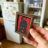 A hand holds a framed picture of a black cat against a red background, intended for refrigerator decor.