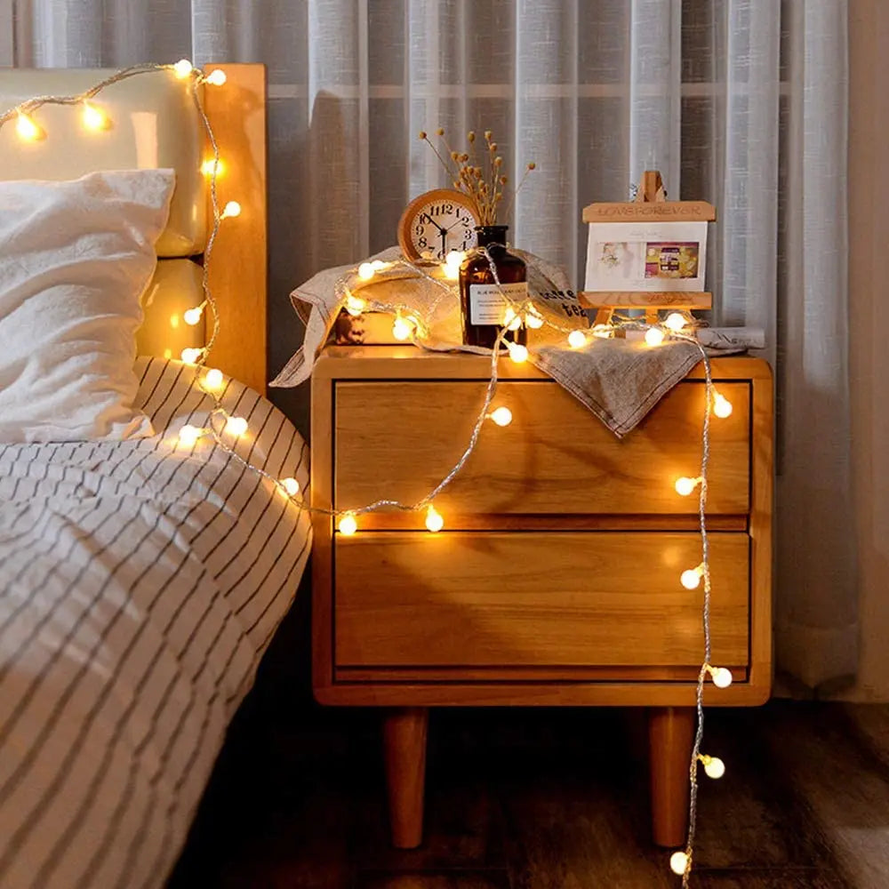 A wooden nightstand decorated with string lights, a clock, and a bottle, next to a bed.