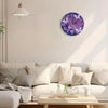 A modern round purple and silver wall clock hangs on a white wall above a light-colored sofa.
