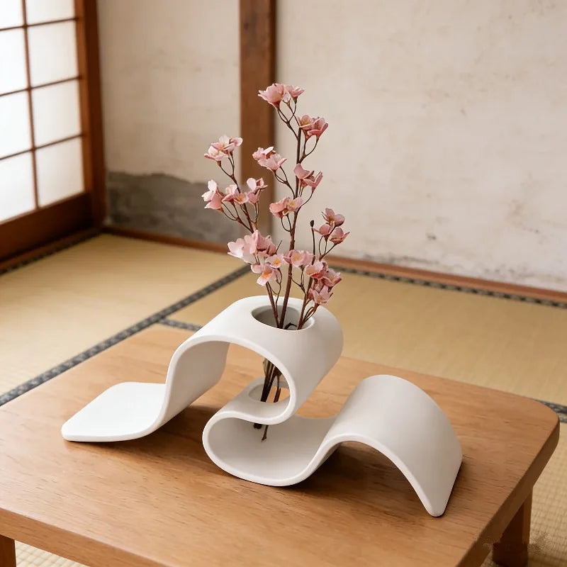 Jarrón moderno curvo blanco con ramas rosadas sobre una mesa de madera.