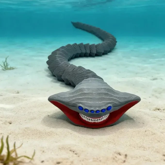 A gray, serpentine sea monster fidget toy with red accents and blue eyes rests on light sand underwater.