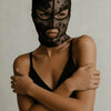 A model wearing a black Venetian Gothic lace mask and a black top, looking directly at the camera.