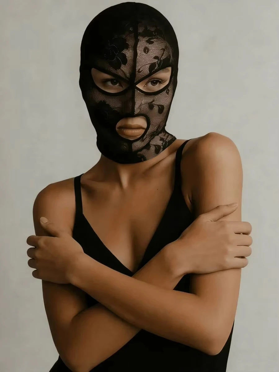A model wearing a black Venetian Gothic lace mask and a black top, looking directly at the camera.