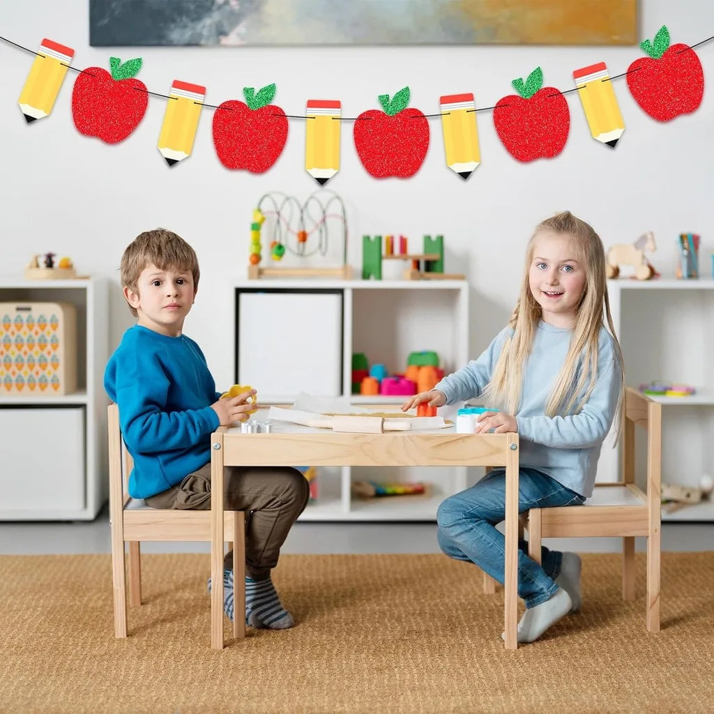 A girl and a boy sitting at a wooden table with a red apple bunting banner above them.