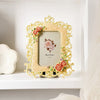 An ornate gold-framed resin photo frame with a light beige mat and floral accents displayed on a white shelf.