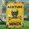 A vintage yellow metal sign with black text warning about mines, displayed outdoors behind a chain-link fence.