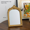 A gold-framed arched carved mirror with a rectangular reflective surface displayed on a wooden surface.