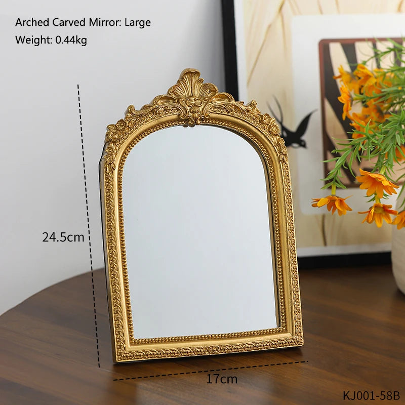 A gold-framed arched carved mirror with a rectangular reflective surface displayed on a wooden surface.
