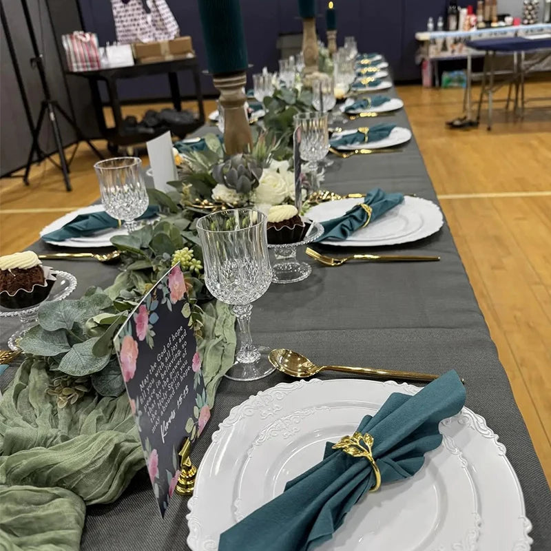 A long table setting display featuring reusable plastic wine glasses, gold cutlery, and dark green foliage.