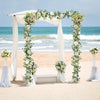 A wedding archway decorated with white fabric and an artificial eucalyptus and rose garland on a sandy beach.