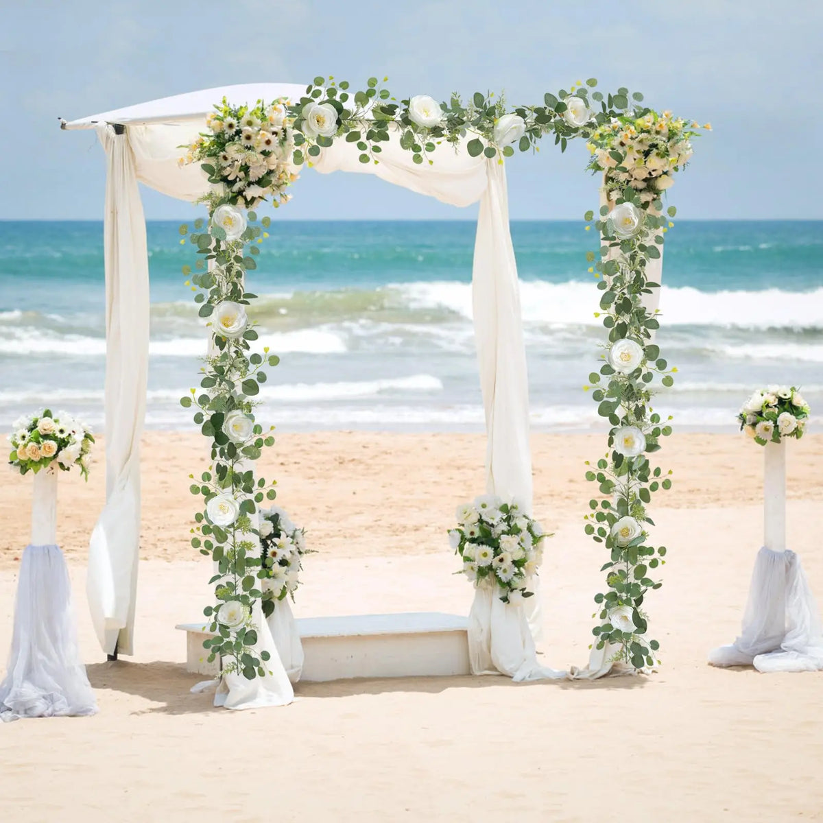 A wedding archway decorated with white fabric and an artificial eucalyptus and rose garland on a sandy beach.