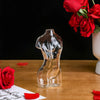 Artistic glass human body vase with a red rose in the background on a wooden surface.