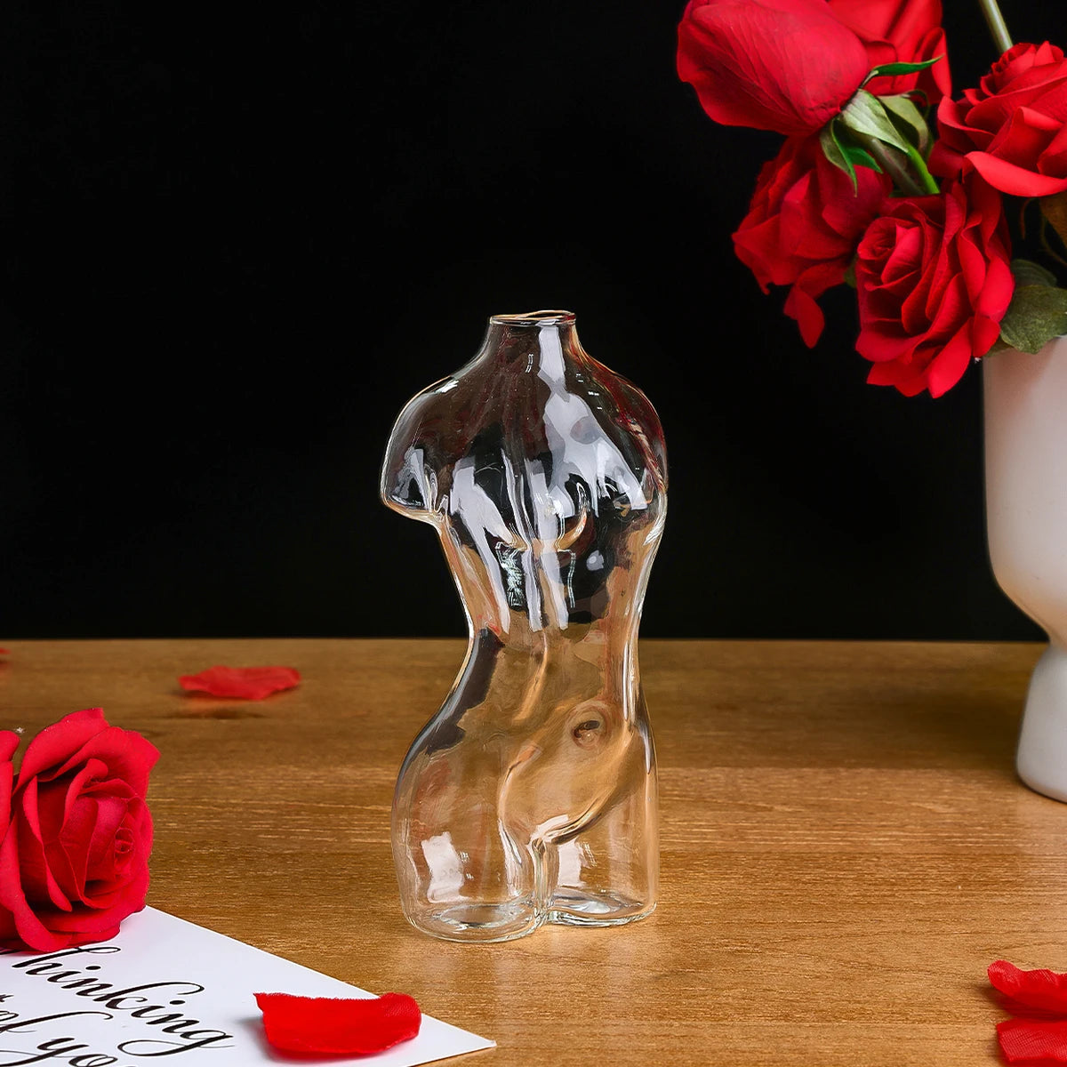 Artistic glass human body vase with a red rose in the background on a wooden surface.