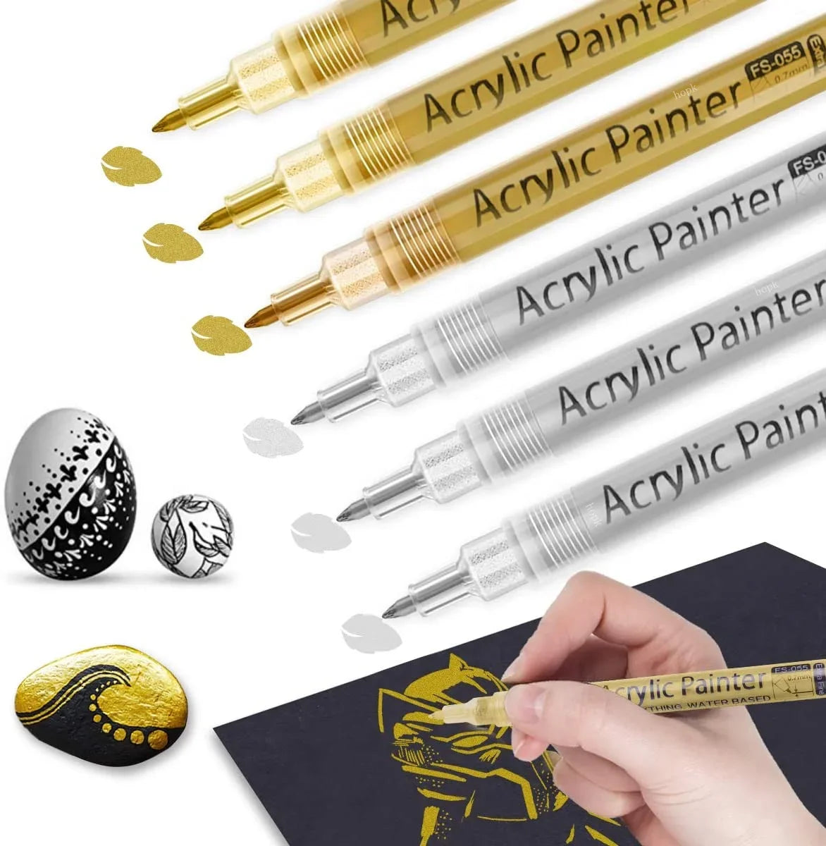 A collection of extra-fine point acrylic paint pens in gold and silver colors are shown with a hand painting on a dark surfac