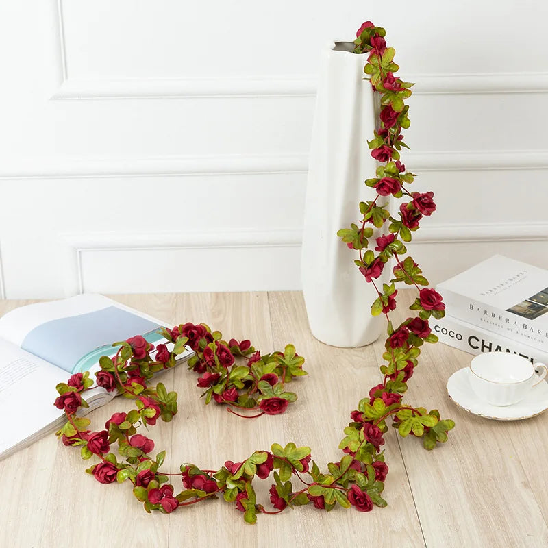 A long garland of deep red roses is displayed in a white vase on a light wooden surface.