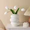 A modern white plastic decorative vase with green leaves holds white tulips on a white table.