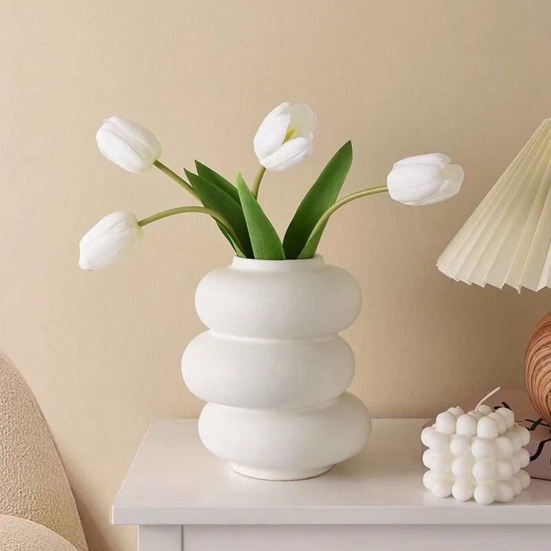 A modern white plastic decorative vase with green leaves holds white tulips on a white table.