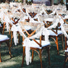 A row of wooden chairs decorated with white lace and fabric runners set up outdoors for a wedding or event.
