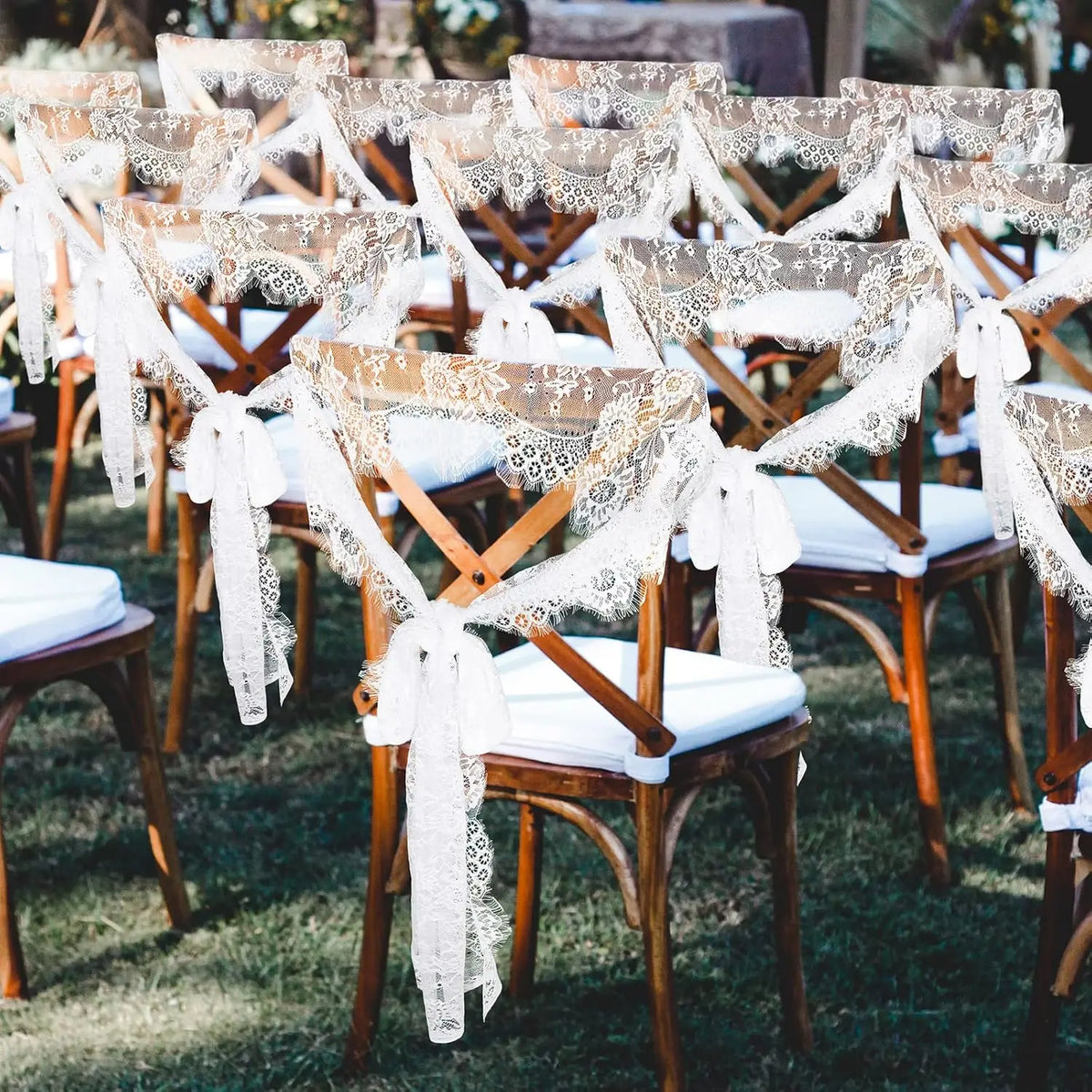 A row of wooden chairs decorated with white lace and fabric runners set up outdoors for a wedding or event.