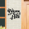 A black metal sign with cursive text welcoming to our home mounted on a white brick wall.