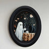 Oval framed wall art depicting a ghost figure and a black cat in a dark floral pattern.