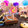A hand holds a wooden stick with decorated lollipop candy shapes on it, surrounded by colorful spherical candies.