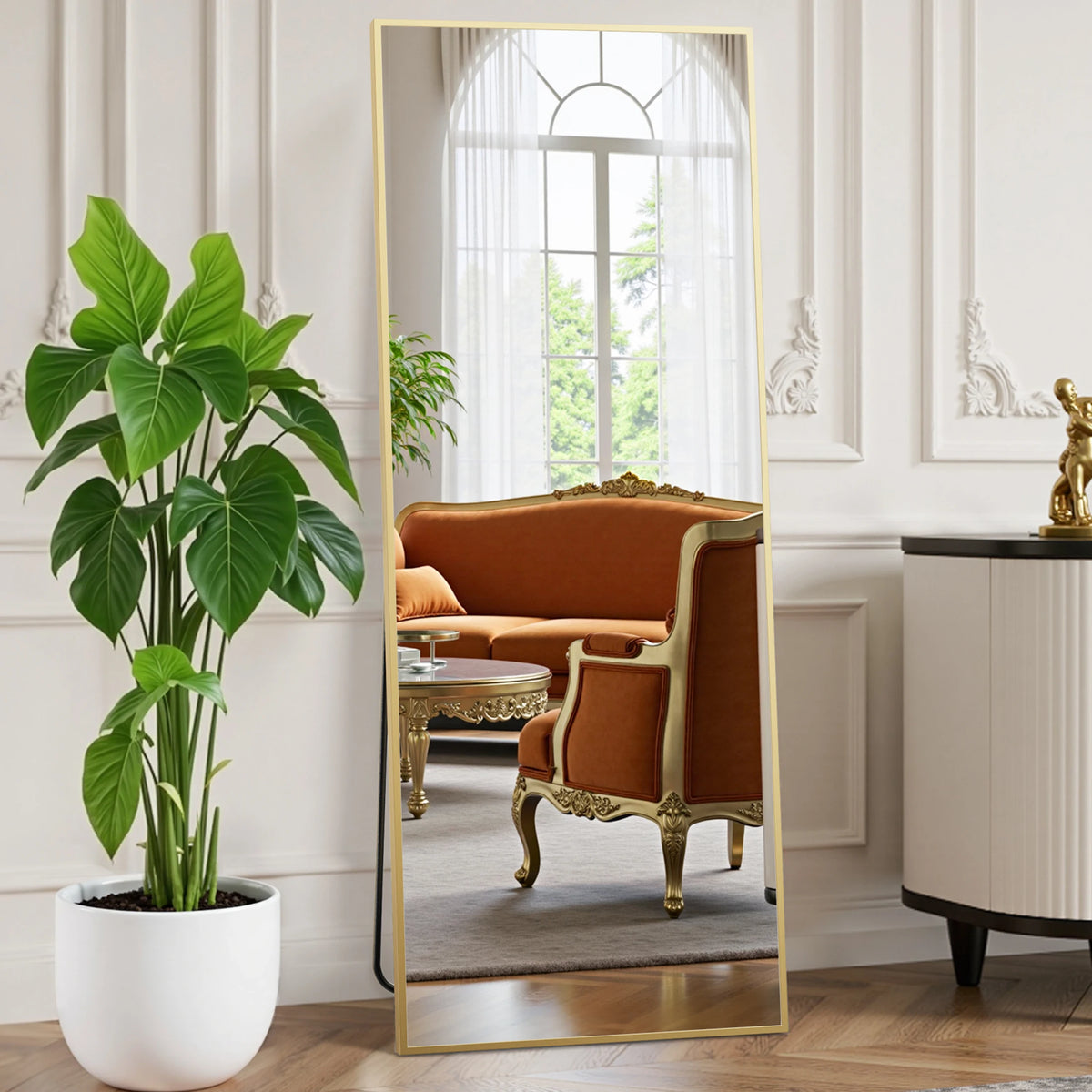 A full length body mirror with a gold frame reflecting an orange velvet sofa and interior decor.