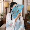 A woman holds a hand-painted decorative folding fan with blue and pink floral designs in an indoor setting.