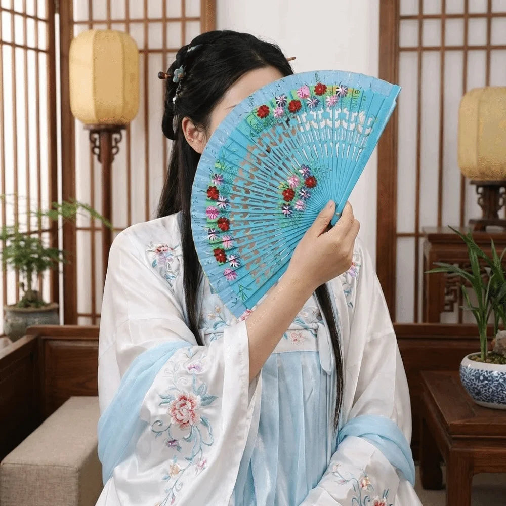 A woman holds a hand-painted decorative folding fan with blue and pink floral designs in an indoor setting.