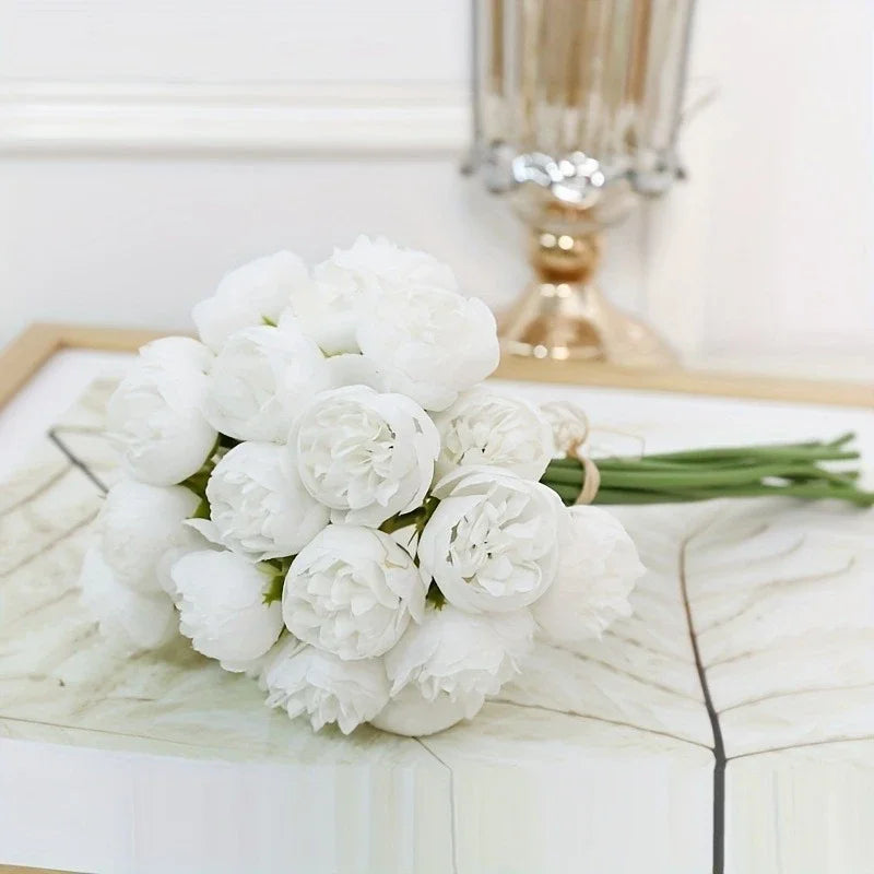 A bouquet of white peonies with green stems displayed on a light surface next to a gold vase.
