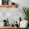A rustic wooden surface displays self-adhesive tile wall stickers in gray and white geometric patterns with greenery.