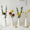 A display of dried greenery and yellow flowers arranged in white ceramic mini vases on a white surface.