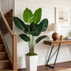 An artificial Bird of Paradise plant in a white planter sits on a wooden table next to a staircase.