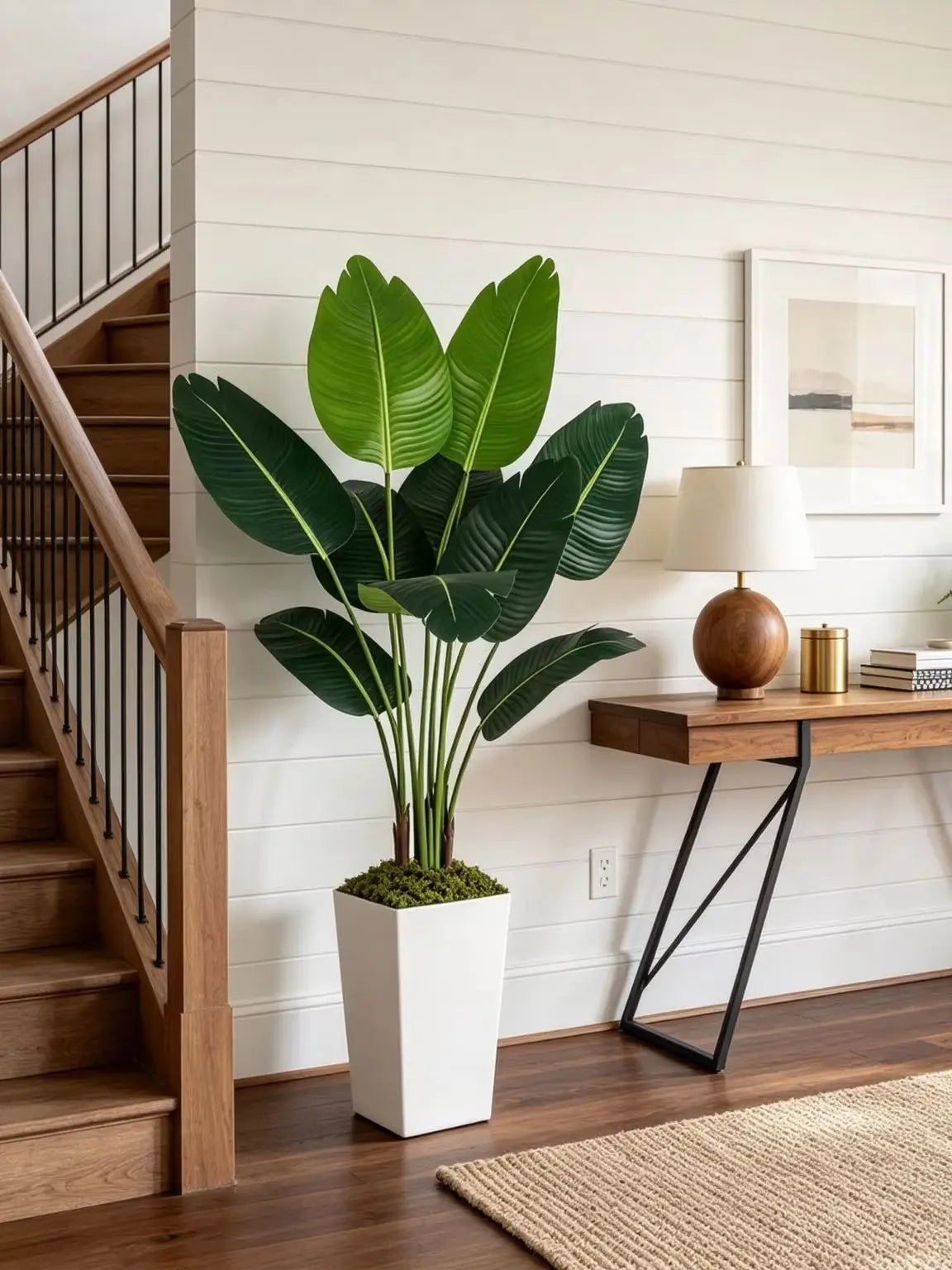 An artificial Bird of Paradise plant in a white planter sits on a wooden table next to a staircase.